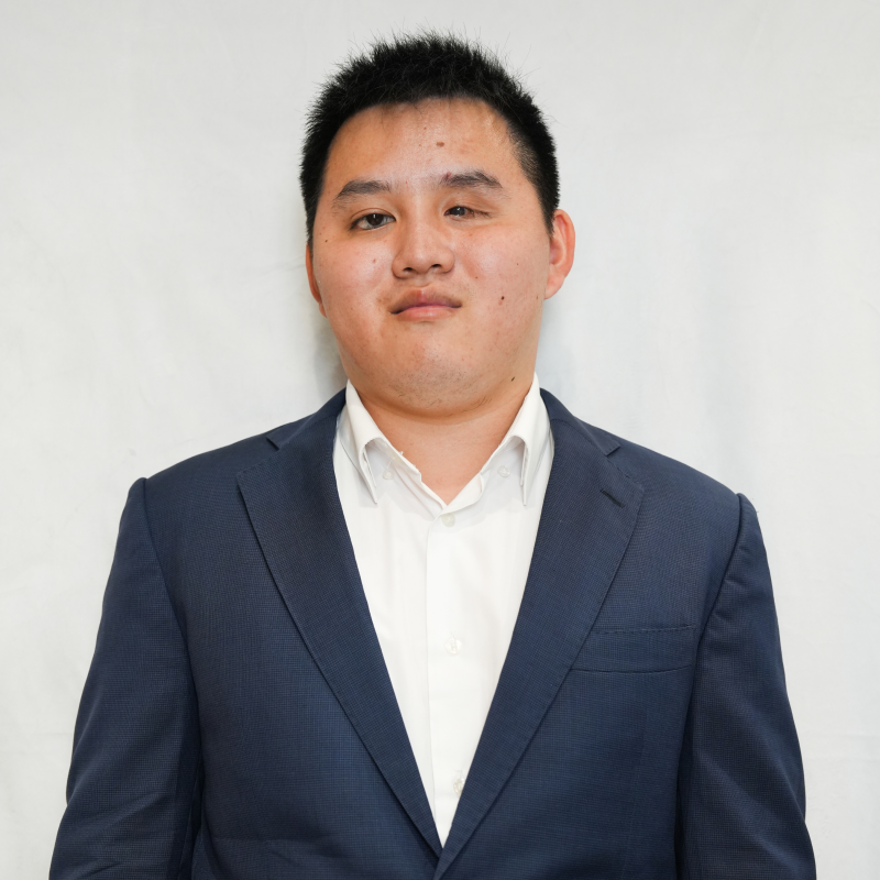 Zhicheng, un jeune homme asiatique vêtu d'un costume bleu marine et d'une chemise blanche, se tient debout devant un fond blanc. Zhicheng sourit et la photographie ressemble à un portrait professionnel.