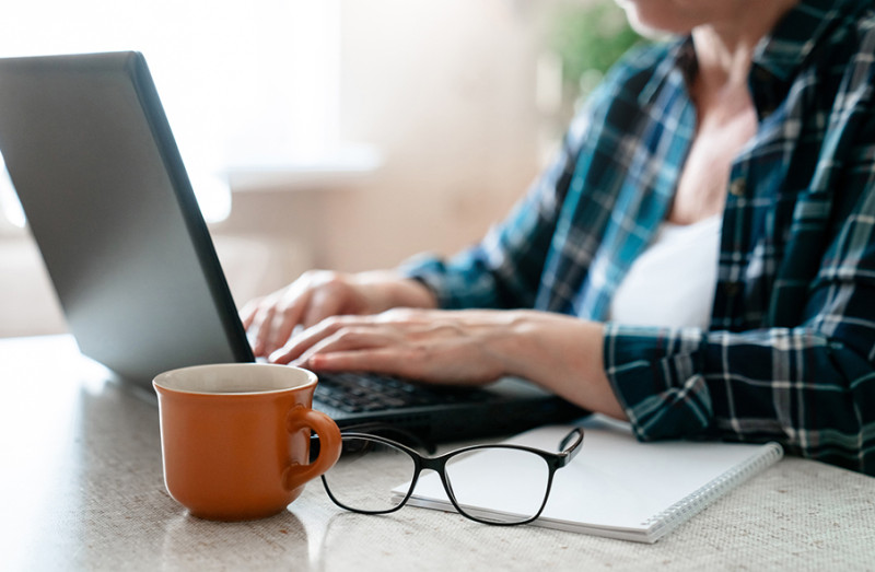 A person with light skin wearing a flannel shirt types on their laptop on a table - a mug, paper, and glasses are nearby