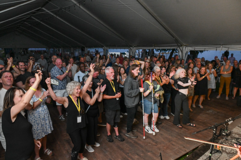 At the Lake Joe Live event, a large crowd of people under a tent cheers, claps, and raises their hands while facing the stage to take in Alan Doyle’s performance.