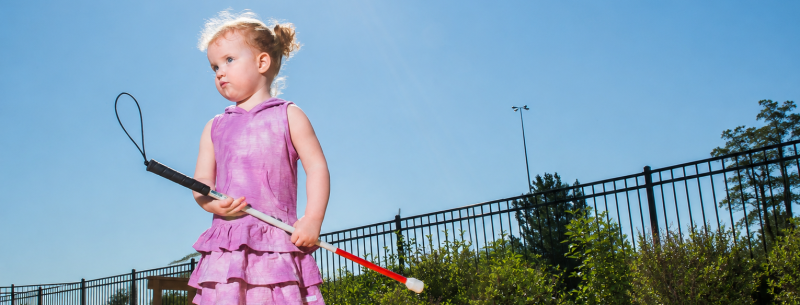 Une jeune fille ayant une perte de vision, vêtue d'une robe rose, tenant une canne blanche à l'extérieur,