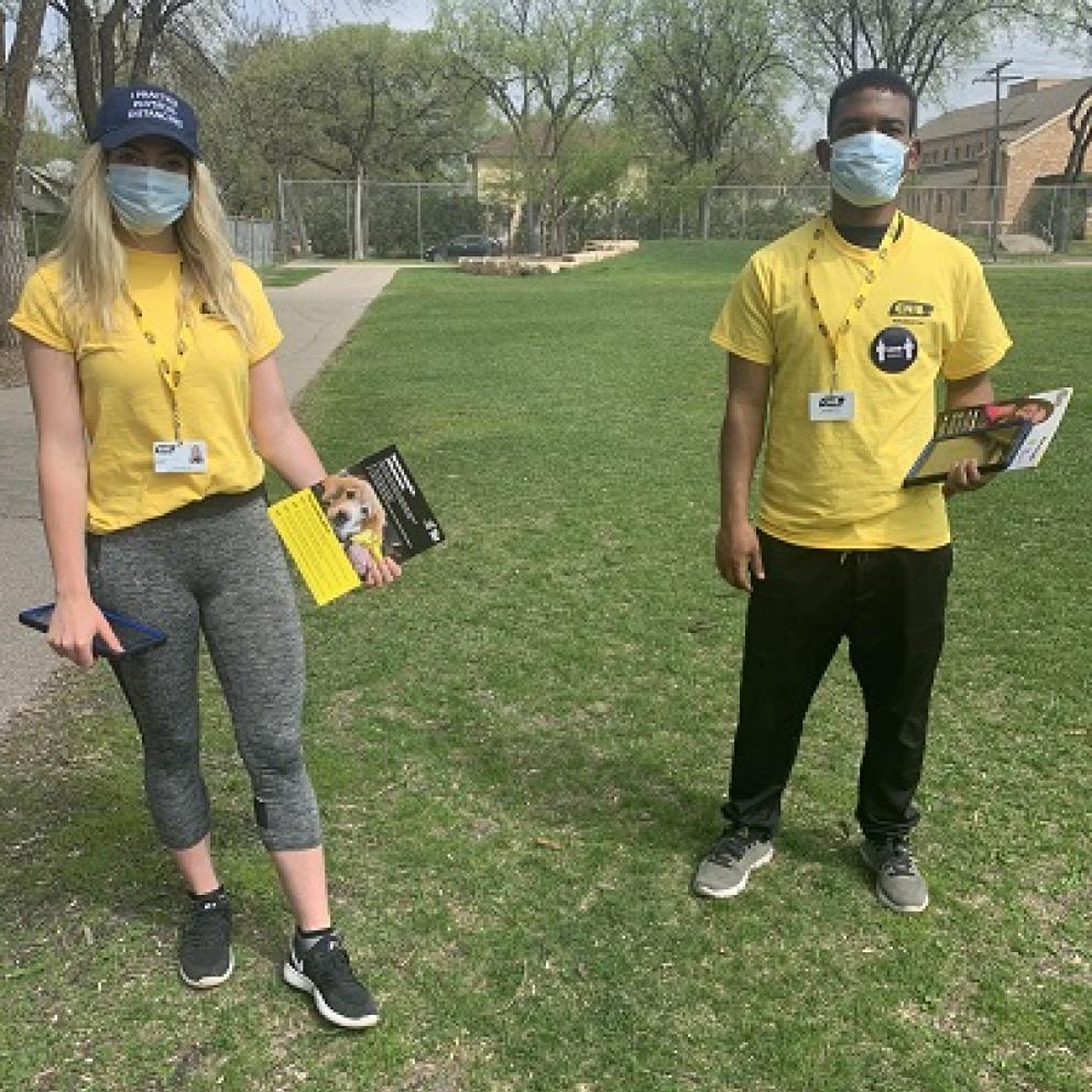 Trois collecteurs de fonds portant des T-shirts jaunes d'INCA et des masques de protection se tiennent à l'écart sur un terrain gazonné, brandissant des brochures de collecte de fonds d'INCA.