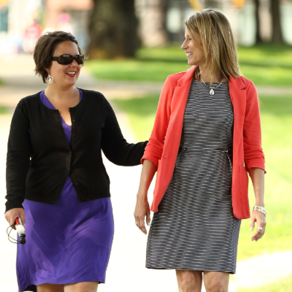 An example of the sighted guide technique. Two woman walk down a sidewalk together. A woman holds the guide's arm lightly above the elbow and allows the guide to walk one-half step ahead.