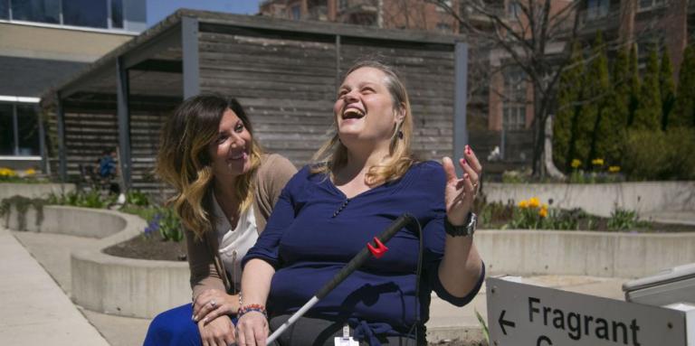 A brunette woman sits and smiles at a blond woman with a white cane, as both sit next to a sign for a fragrant garden.