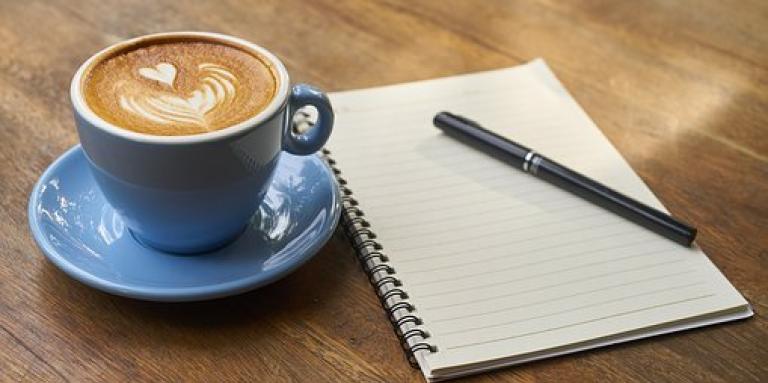 Une tasse de café latté avec un cœur formé dans la mousse, déposée sur une table en bois, à côté d'un pad de note.