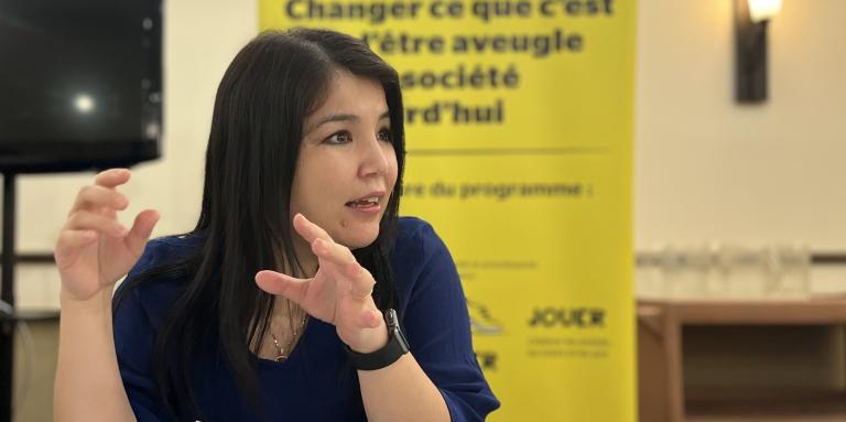 Najla parle avec ses mains, assise,  à un table, devant la bannière Changer ce que signifie être aveugle d'INCA.