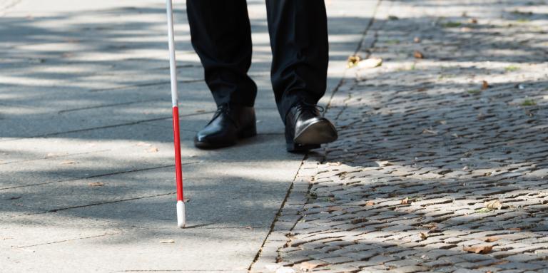 Une canne blanche balaie un trottoir. En arrière-plan, des chaussures d'homme suivent le bout de la canne