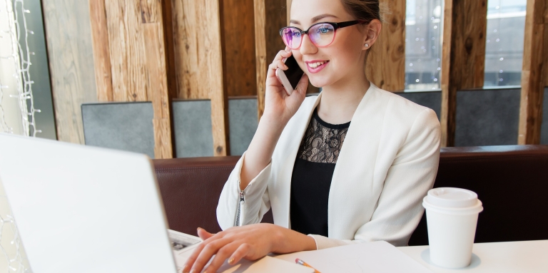Une jeune femme  portant des lunettes parle au téléphone et est sur l'ordi. un café pour emporté est déposé sur la table. 