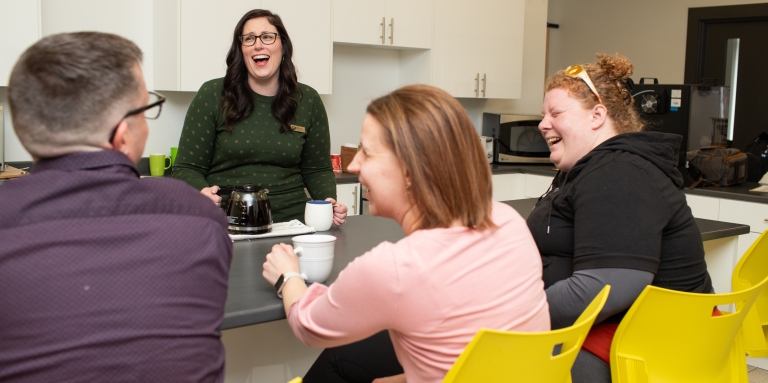 3 jeunes femmes et un jeune homme rient de bon coeur autour d'un bon café dans une cuisine.