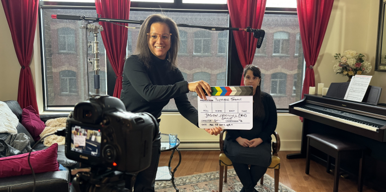 Une femme souriante qui tient le clap cinéma, on aperçoit une camera en avant plan et en arrière plan une femme assis sur une chaise