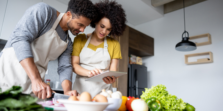 Un jeune homme et une jeune femme portant un tablier, consultent une recette sur une tablette.  Le jeune homme coupe une partie des ingrédients qui sont devant eux.