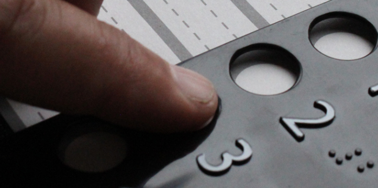 Gros plan sur une main qui explore au toucher un gabarit de vote tactile en relief, avec des chiffres 1, 2, 3 et leurs équivalents en braille, posé sur un bulletin de vote.