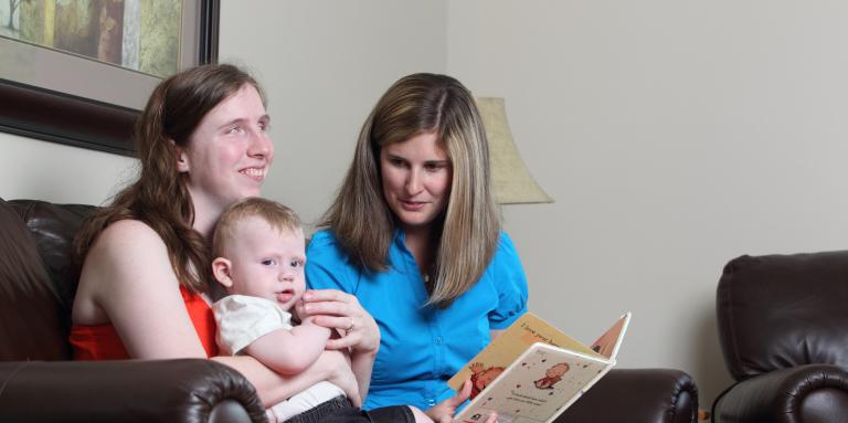 Two women sit on a couch. The woman on the left is holding an infant while the other woman is holding a storybook.