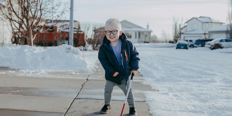 Little boy on a sidewalk with snow around him, holding his white cane like a hockey stick. He's smiling.