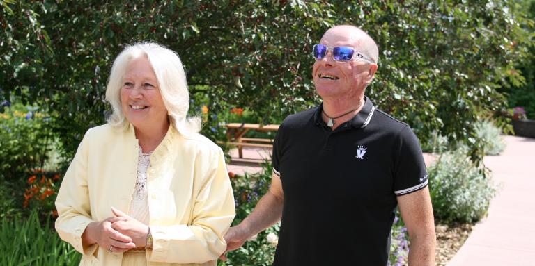 A woman in yellow guides a man in a black t-shirt. The man is holding on to her elbow. Both are smiling.