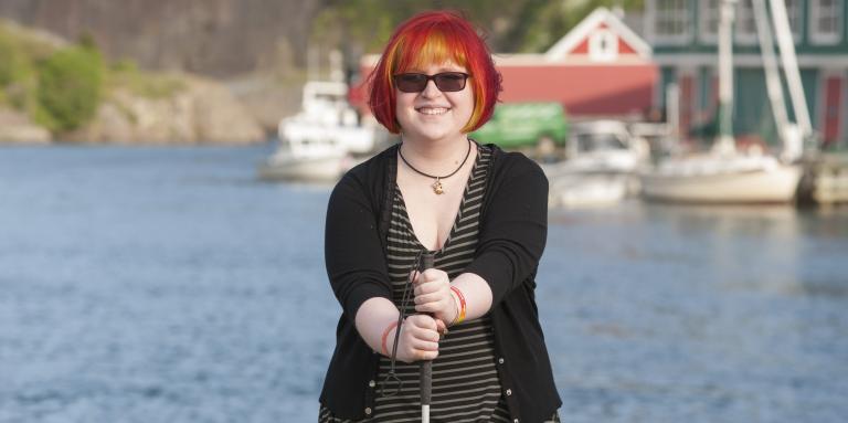 Young woman standing in front of a body of water with boats behind her, smiling at the camera holding her white cane in front of her.