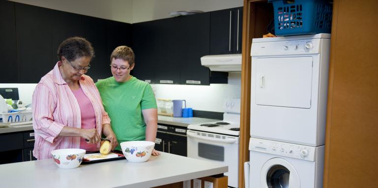 Two women are standing in front of a counter with a potato, cutting board and two bowls. The woman on the left in pink is peeling the potato, while the woman on the right, wearing green, if overseeing.