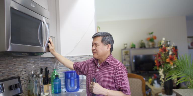 Man in a red plaid shirt is holding his white cane to his side while reaching for the microwave.