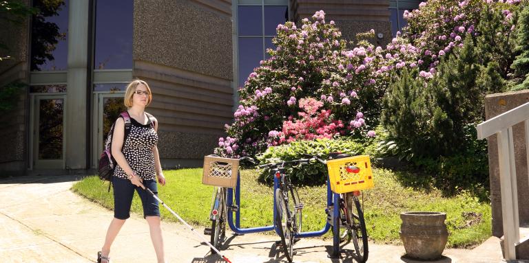 Young woman walking with her white cane on a sidewalk passing by three parked bicycles.