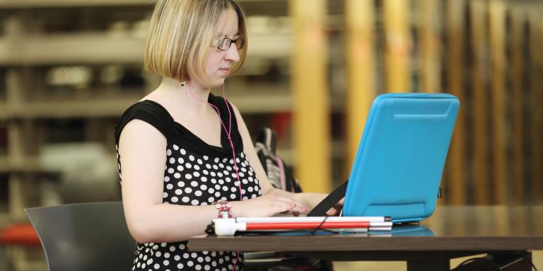 A woman sits at a desk with her laptop open and her collapsed white cane next to it.