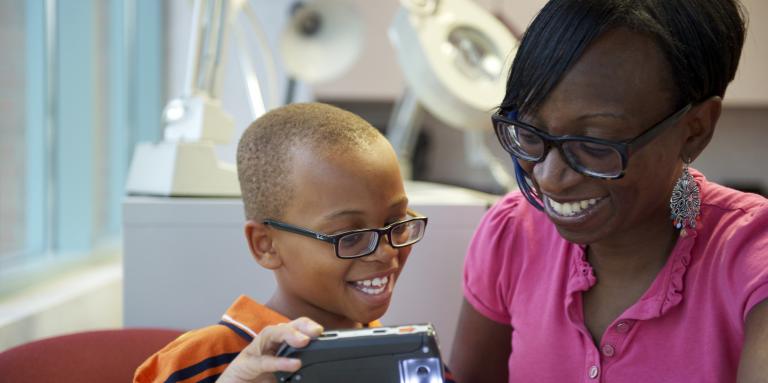 A young boy and his mother, both wearing glasses, interact as the little boy plays with a magnifier. 