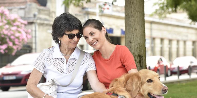 Two women sitting at a bench. The woman on the right has a white cane next to her, and the woman on the left has a golden labrador guide dog.