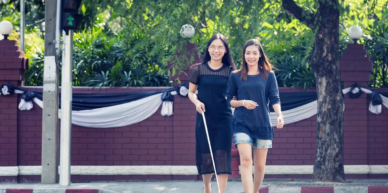 Deux femme traversant la rue. Une utilise une canne blanche et l'autre la guide.