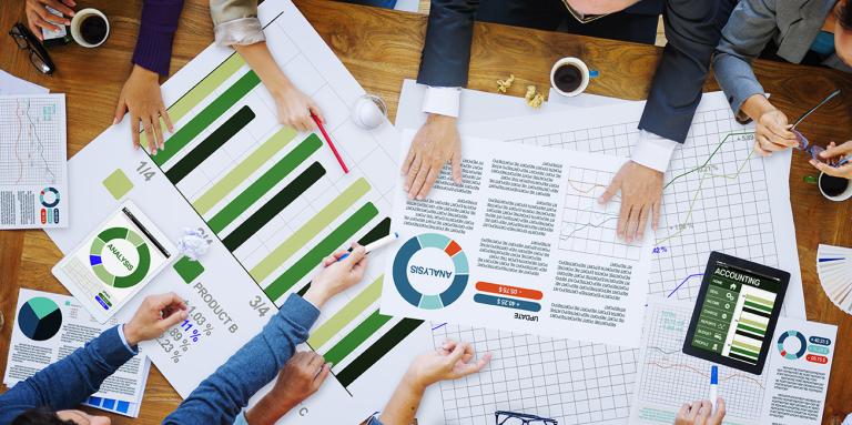 an overhead view of six people sitting around a table working on some research documents which include graphs and tables.