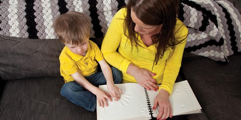 Un petit garçon assis à côté de sa mère sur un sofa lit un livre tactile.