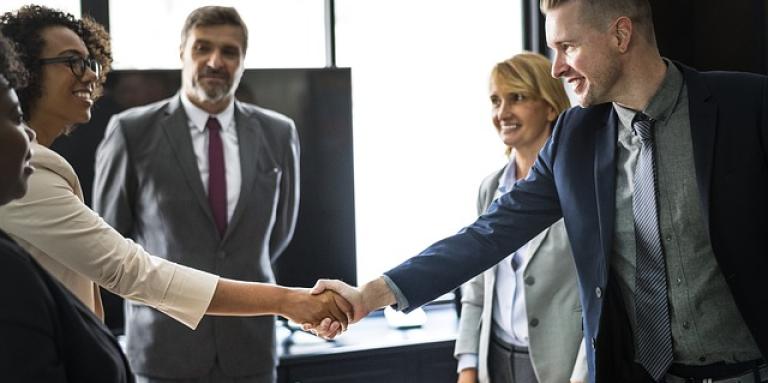 The closing of a presentation: Three women and two men stand around a table. A man and woman shake hands.
