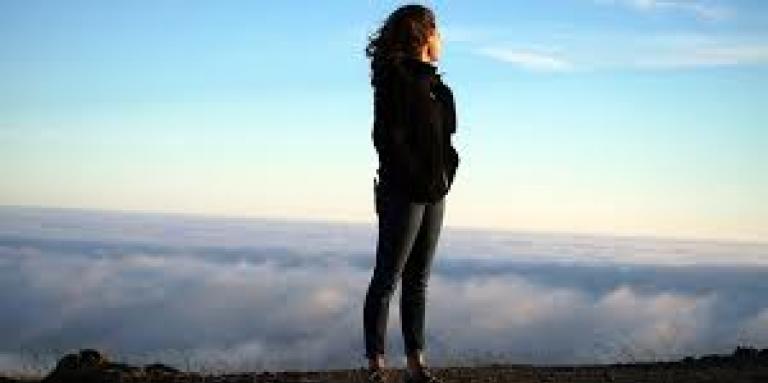A woman stands on top of a mountain, looking out over the clouds and ground below. Her hair blows in the breeze and the sky is bright blue.