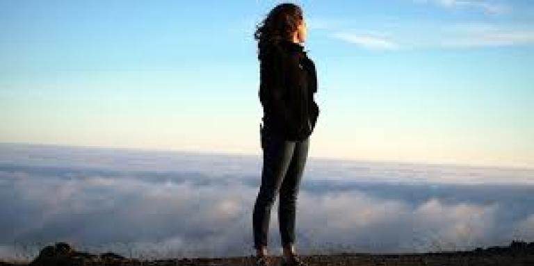 A woman stands on top of a mountain, looking out over the clouds and ground below. Her hair blows in the breeze and the sky is bright blue.