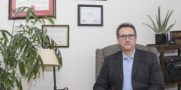 Joshua Simmonds sits in a chair in his office, wearing a suit. There are certificates on the wall behind him, and a coffee table and plants on the left side.