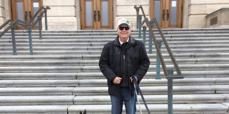 Bob Huber stands outside of the Saskatchewan legislative building with his two dogs, smiling with his sunglasses on and green Roughrider ball cap.