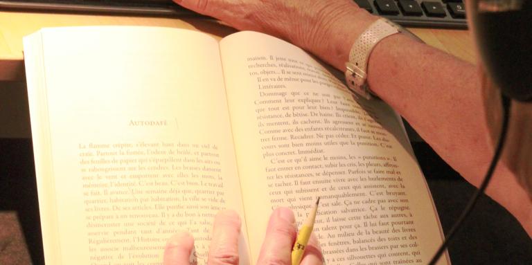 Gros plan d'une femme devant un livre, crayon à la main, écouteur sur les oreille et sa seconde main sur un clavier devant des outils d'enregistrement