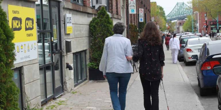 Une bénévole et une jeune femme aveugle marche en face du bureau d'INCA on voit le pont Jacques-Cartier à l'horizon