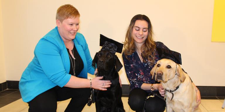 Ashley et Danika accompagnées de deux chiens Labrador/Golden Retriever croisés (un noir et un jaune) portant des chapeaux de graduation.