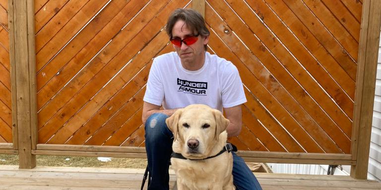 Gary Peasgood, standing outdoors against a wood patio wall with his guide dog. 