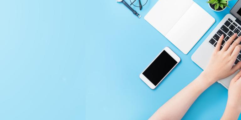 Deux mains qui tapent sur le clavier d'un ordinateur portable. On voit un iPhone sur la table bleue à côté de l’ordinateur.