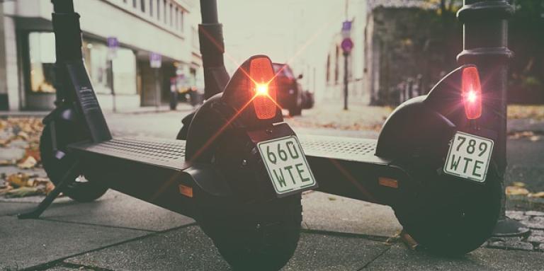 Deux trottinettes électriques l’une à côté de l’autre sur un trottoir. Les feux de freinage clignotent en rouge.