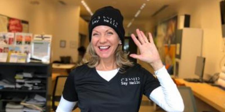 Denise, an entrepreneur, wears her "Say Hello 2 Blindness" attire and waves at the camera with her left hand. 