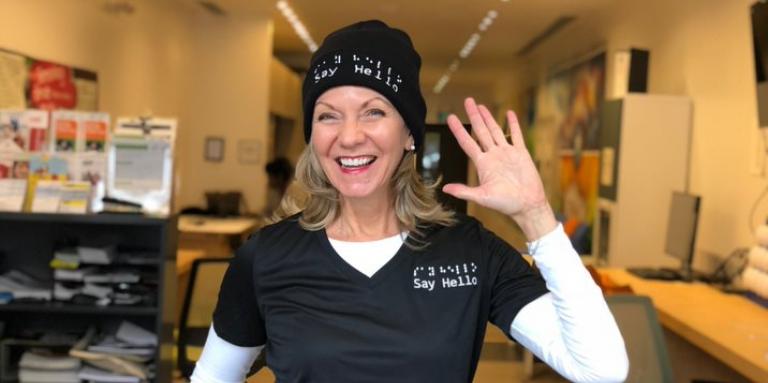 Say Hello 2 Blindness founder Denise Justin smiles and waves while wearing her company-branded shirt and toque.