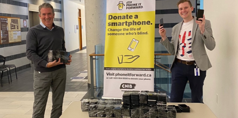 John M. Rafferty et Thomas Simpson, souriants, sont debout à une table et tiennent plusieurs téléphones intelligents donnés par la Banque du Canada. Sur la table sont également empilés des dons de téléphones intelligents. Derrière eux se trouve un présentoir portant la bannière du programme Rendre l'appareil.