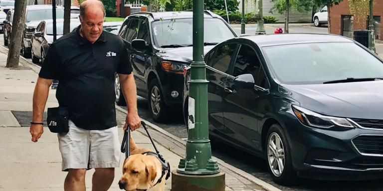 Carl marchant sur un trottoir avec un chien-guide d'INCA en formation.]