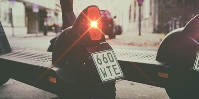 A photo of two e-scooters parked on a sidewalk, both with red tail lights and license plates. 