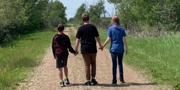 The backs of Ashley’s three children as they walk down a secluded road in a lush, green area. The children are holding hands.