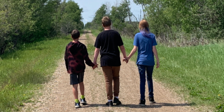 Les trois enfants d'Ashley vus de dos alors qu'ils marchent le long d'une route isolée dans une zone verte et luxuriante. Les enfants se tiennent la main.