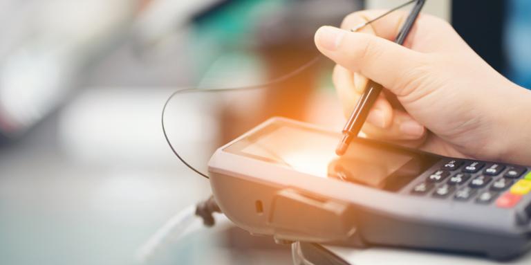 A close up of a person’s hand using a stylus pen to make a selection on the touch screen surface of a payment terminal.