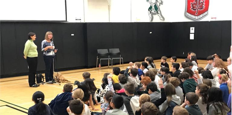 Two Community Engagement Volunteers deliver a presentation in front of a large group of school-aged children sitting in a gymnasium.