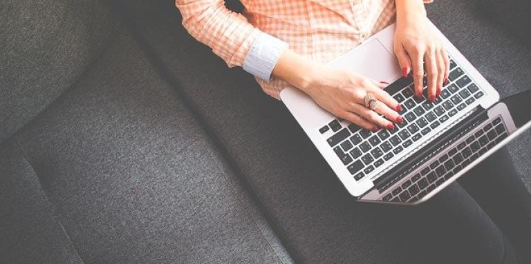 The upper torso of a woman as she sits on a couch and types on a laptop.