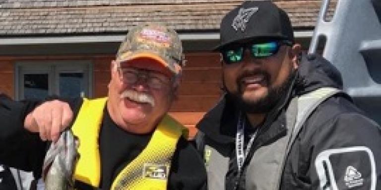 Bruce Roulston posing with a caught fish on a dock beside Eugene Chong at CNIB Lake Joe. They are both wearing life jackets and a ballcap.